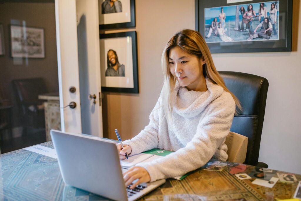 Asian woman in a white sweater working remotely on a laptop at home.